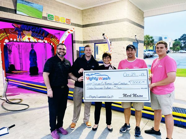 Group in pink shirts holding a $7,000 Mighty Wash check for Susan G. Komen South Carolina at the car wash tunnel entrance
